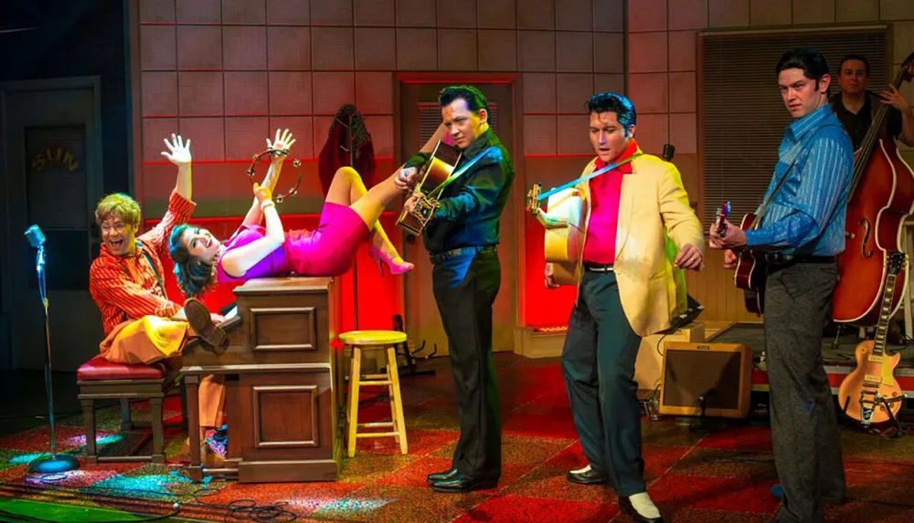 The image shows a lively theatrical performance with musicians in 1950s attire and a woman playfully posed on a desk evoking a vibrant rock n roll scene
