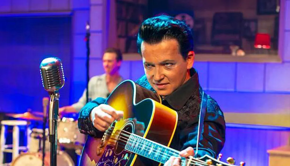 A man dressed in a vintage-style outfit is intensely playing an acoustic guitar near a classic microphone with a drummer blurred in the background