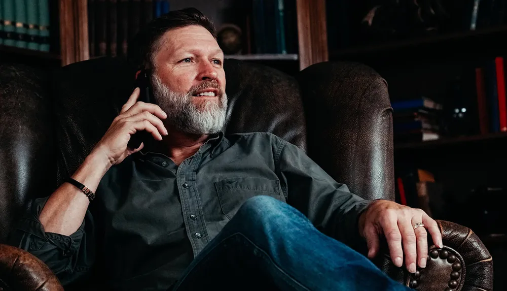 A man with a beard is relaxed in a leather chair smiling as he talks on a smartphone