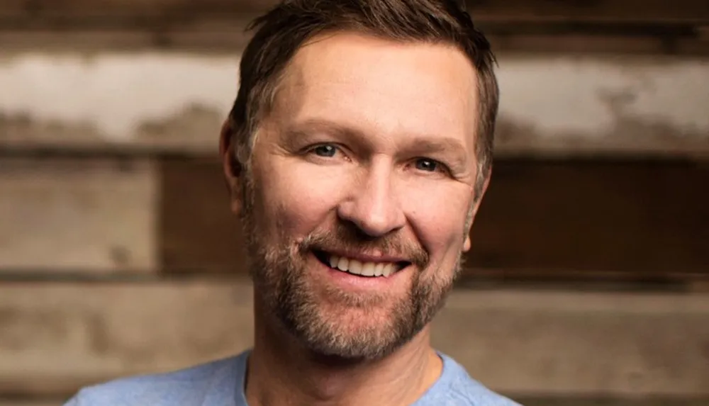 The image shows a smiling man with stubble and short hair against a blurred wooden backdrop