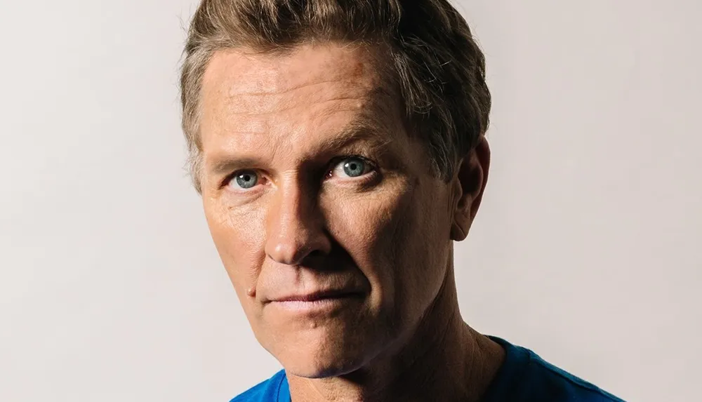 The image shows a close-up of a mature man with striking blue eyes wearing a blue shirt and giving a serious look towards the camera