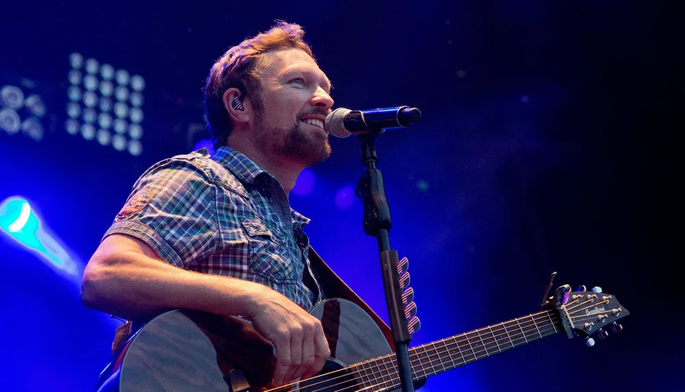 A male musician with a beard is performing live singing into a microphone with an acoustic guitar in his hands surrounded by colorful stage lighting