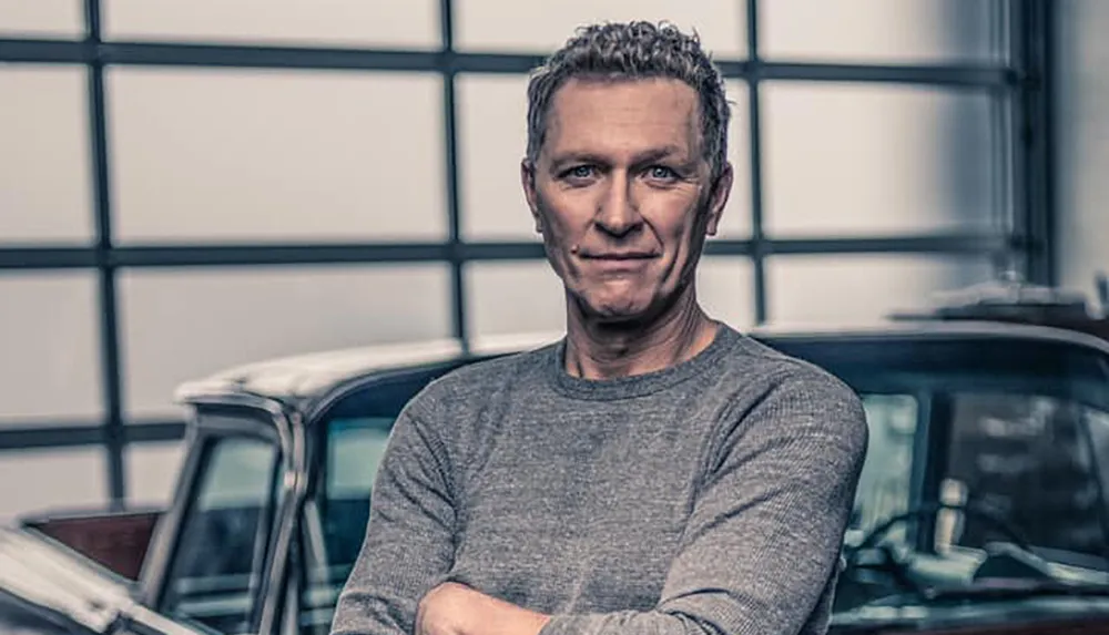 A man with crossed arms is standing in front of a vintage car inside a garage with large windows in the background