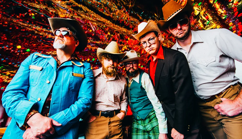 Five men posing confidently in eclectic and colorful cowboy-inspired attire stand in front of a vibrant festive backdrop