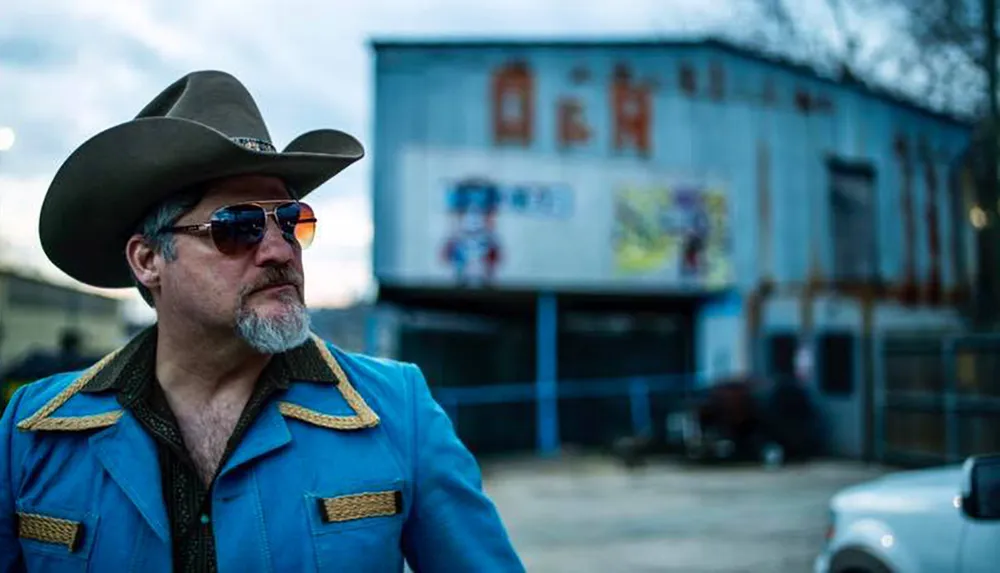 A man in a cowboy hat and blue western-style jacket stands in the foreground with an industrial-looking building blurred in the background