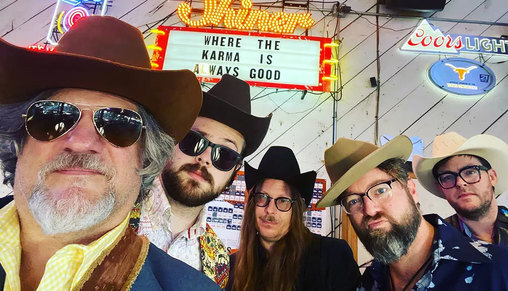 A group of five men wearing cowboy hats pose for a selfie with a neon sign stating Where the Karma Is Always Good in the background