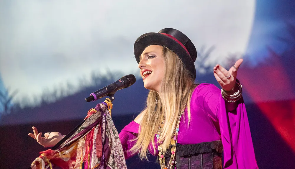 A performer in a vibrant purple dress and a black hat is singing into a microphone with an expressive gesture against a blurred background that appears to be a stage setting