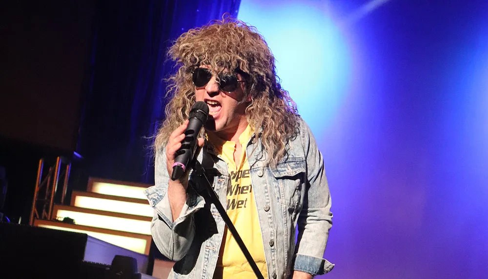 A person with curly blonde hair and sunglasses is energetically singing into a microphone on a stage with blue lighting in the background