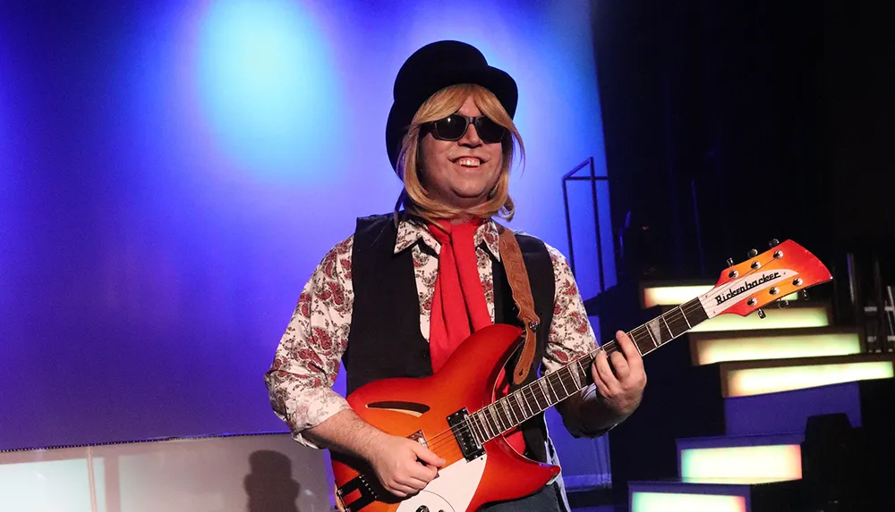 A smiling person wearing sunglasses a black hat and a vibrant shirt is playing a red and white electric guitar onstage with colorful lighting in the background