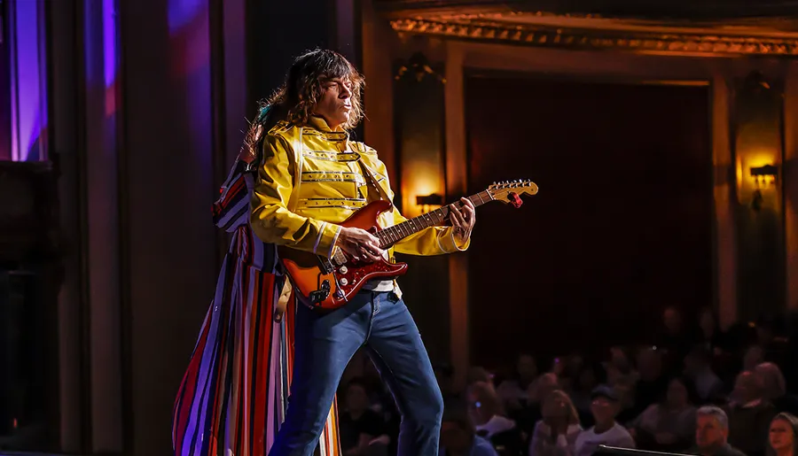 A musician in a yellow jacket plays an electric guitar on stage in front of an audience.