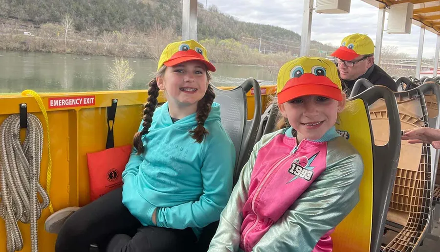 Two smiling children are wearing duck-themed hats while seated on a yellow vehicle that appears to be designed for tours, with a body of water and natural scenery in the background.