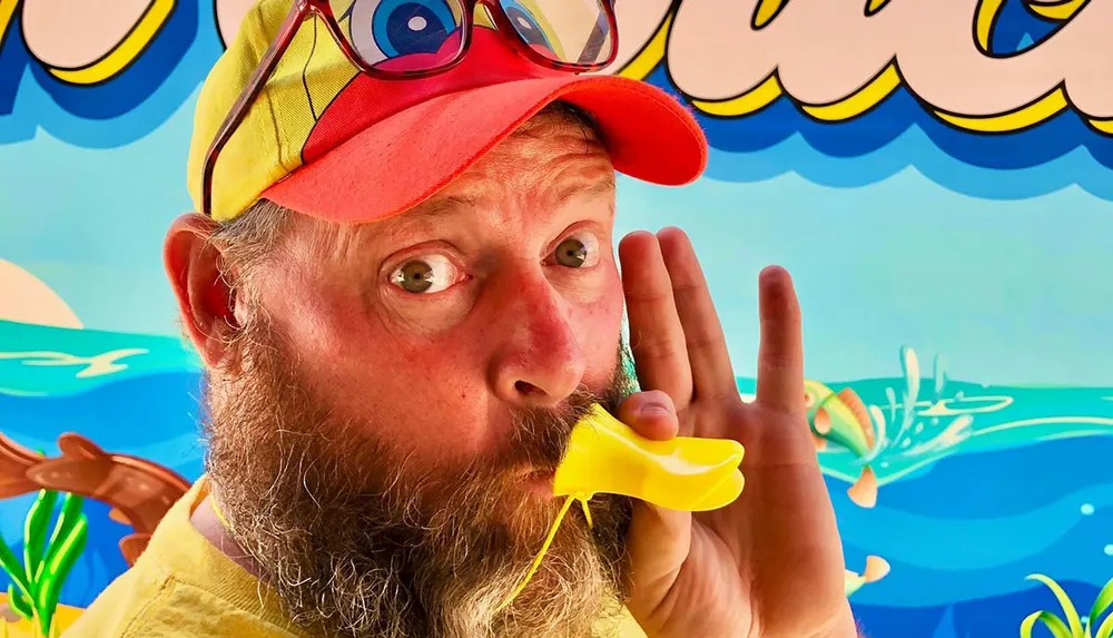 A person with a beard is playfully posing with a yellow duck-shaped whistle in his mouth against a cartoonish aquatic backdrop