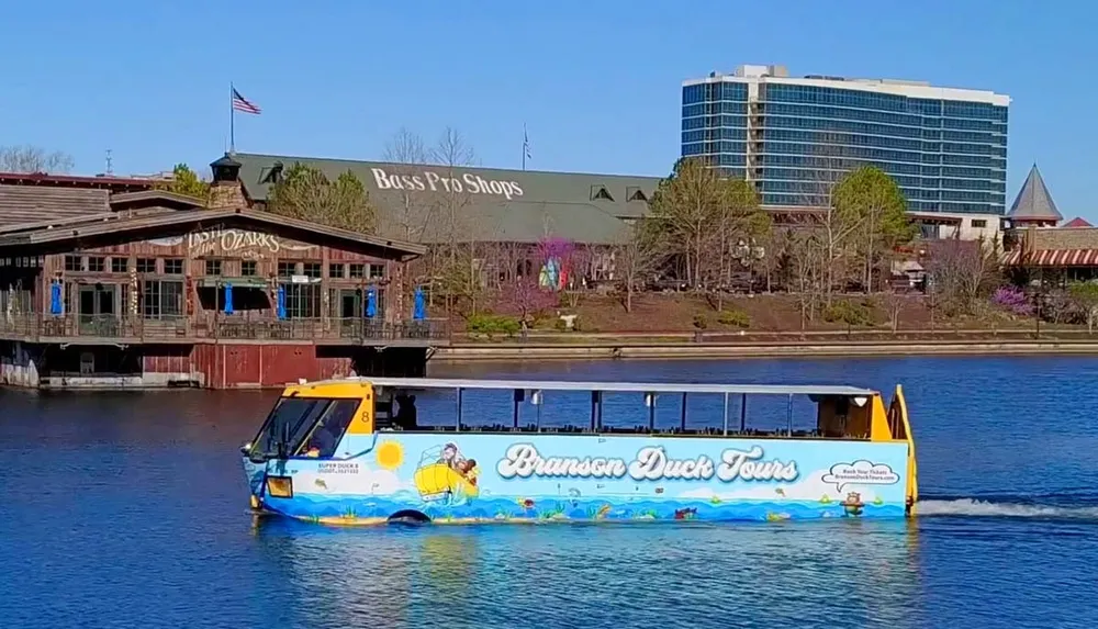 The image shows a colorful amphibious tour vehicle known as a duck boat cruising on a body of water with buildings including a Bass Pro Shops store in the background