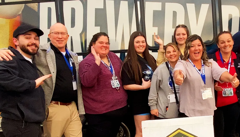 A group of eight smiling people with lanyards are posing for a photo in front of a backdrop with the word BREWERY on it