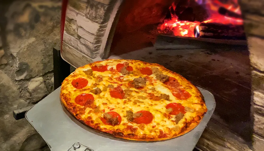 A freshly baked pizza with pepperoni and sausage toppings is sitting in front of a fiery wood-fired oven