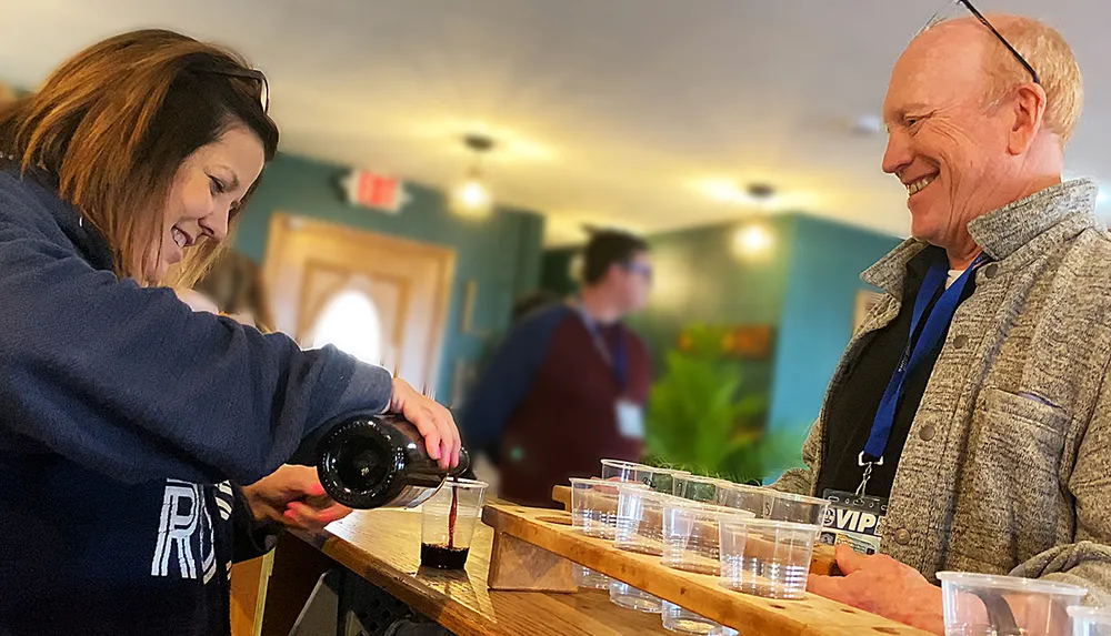 A smiling woman is pouring a beverage into cups for a cheerful man at an indoor event