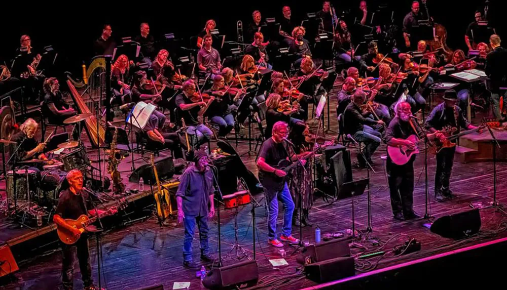 A band performs on stage in front of an orchestra suggesting a fusion of rock and classical music styles in a concert setting