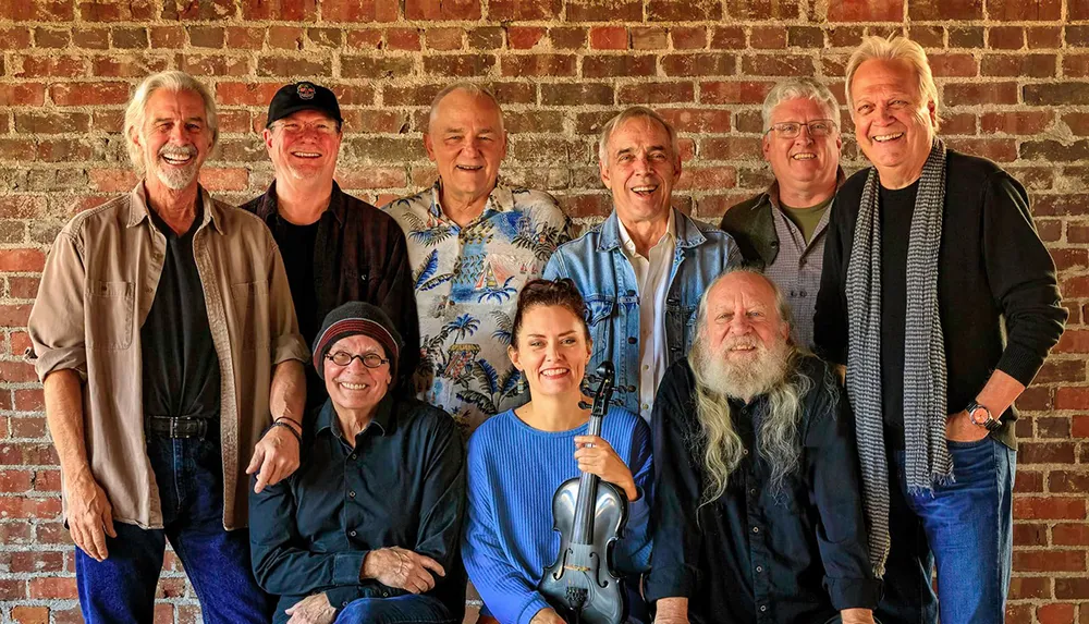 A group of smiling people including men and one woman holding a violin pose together against a brick wall backdrop