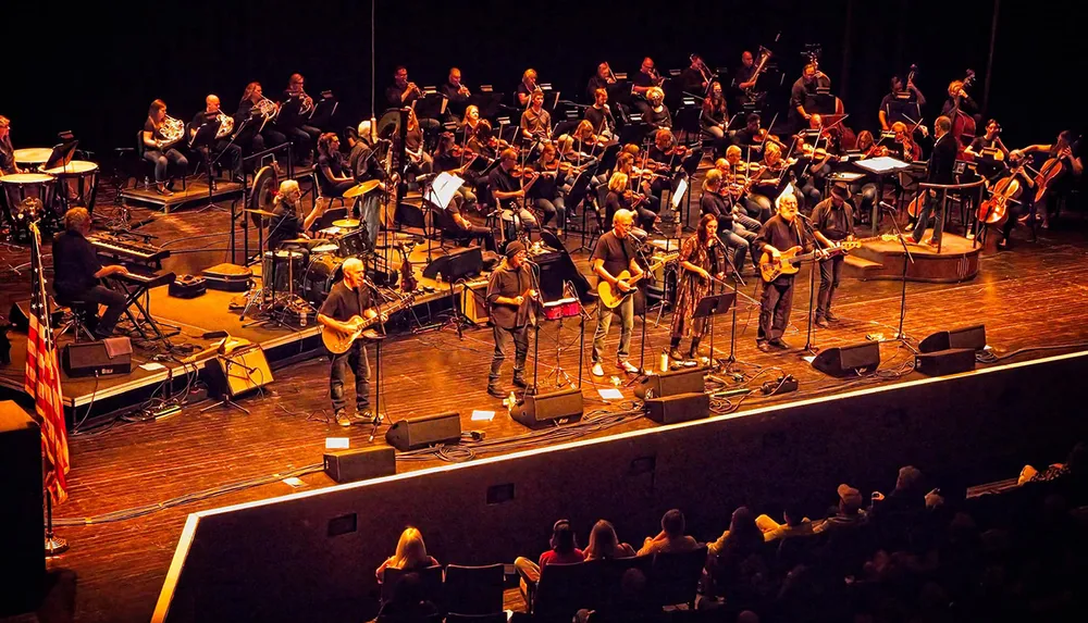 The image shows a vibrant concert with a large ensemble of musicians on stage including a rock band and an orchestra performing for an audience