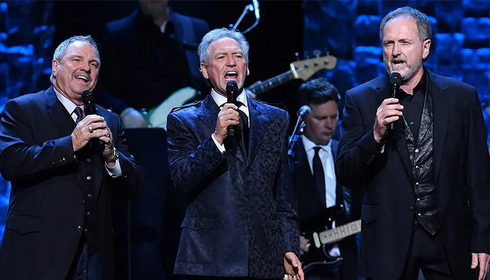 Three men are singing into microphones on stage likely during a live performance with musicians in the background