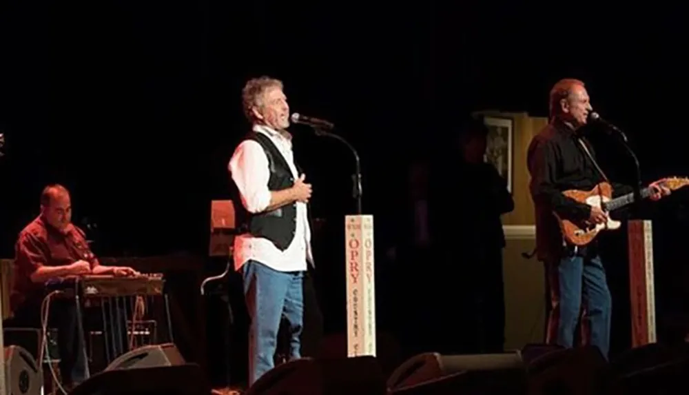 Musicians perform on stage with a singer at the center a steel guitar player seated to the left and a guitarist to the right in front of a microphone stand labeled OPRY