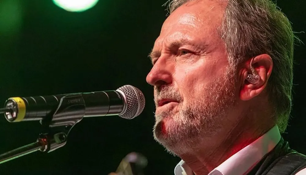 A mature male musician is performing live singing into a microphone on stage under colored lighting