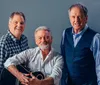 Three smiling men one holding a guitar pose together for a friendly portrait against a gray background