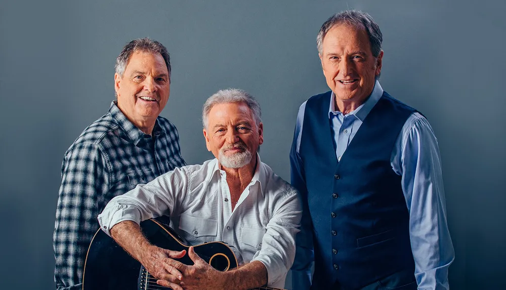 Three smiling men one holding a guitar pose together for a friendly portrait against a gray background