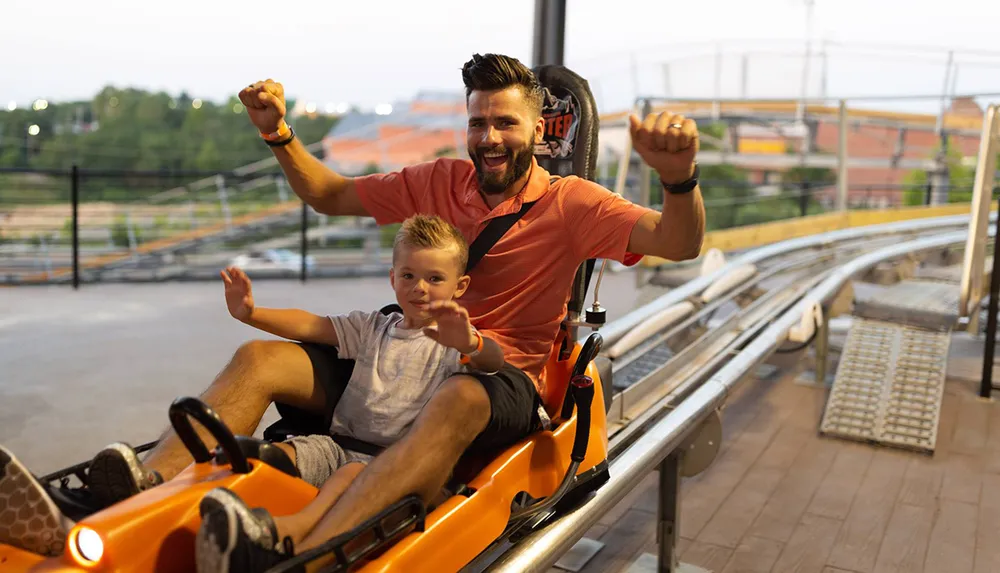 A man and a young boy are raising their fists in excitement while seated in an orange roller coaster cart
