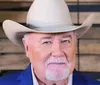 A man with a white mustache and beard is wearing a large white cowboy hat and a blue jacket posing in front of a wooden background