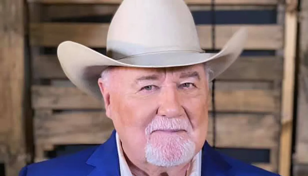 A man with a white mustache and beard is wearing a large white cowboy hat and a blue jacket posing in front of a wooden background