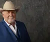 A man with a white mustache and beard is wearing a large white cowboy hat and a blue jacket posing in front of a wooden background