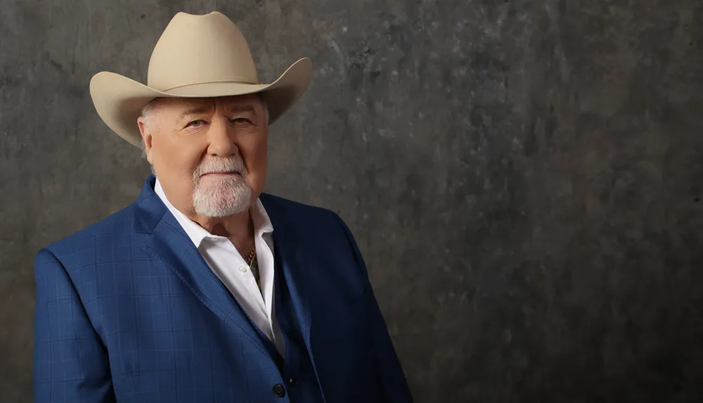 A man in a cowboy hat and blue blazer poses confidently against a textured grey background