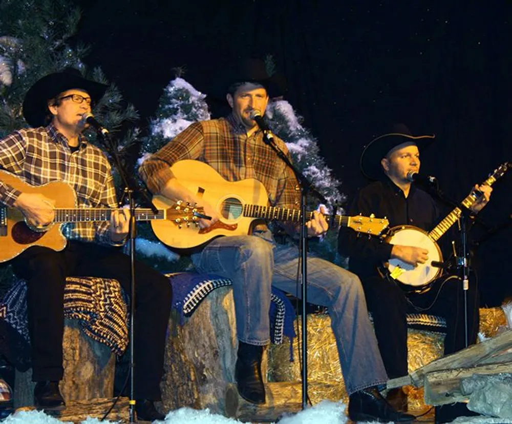 Three musicians wearing cowboy hats are seated on stage playing acoustic guitar and banjo with snowy pine trees as a backdrop creating a cozy winter concert scene
