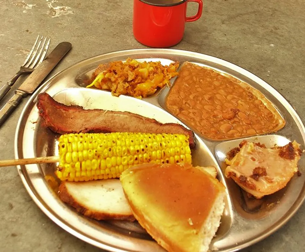 The image shows a hearty meal consisting of baked beans corn on the cob a slice of brisket rolls and what appears to be a portion of a casserole served on a metal tray with utensils and a red mug on the side
