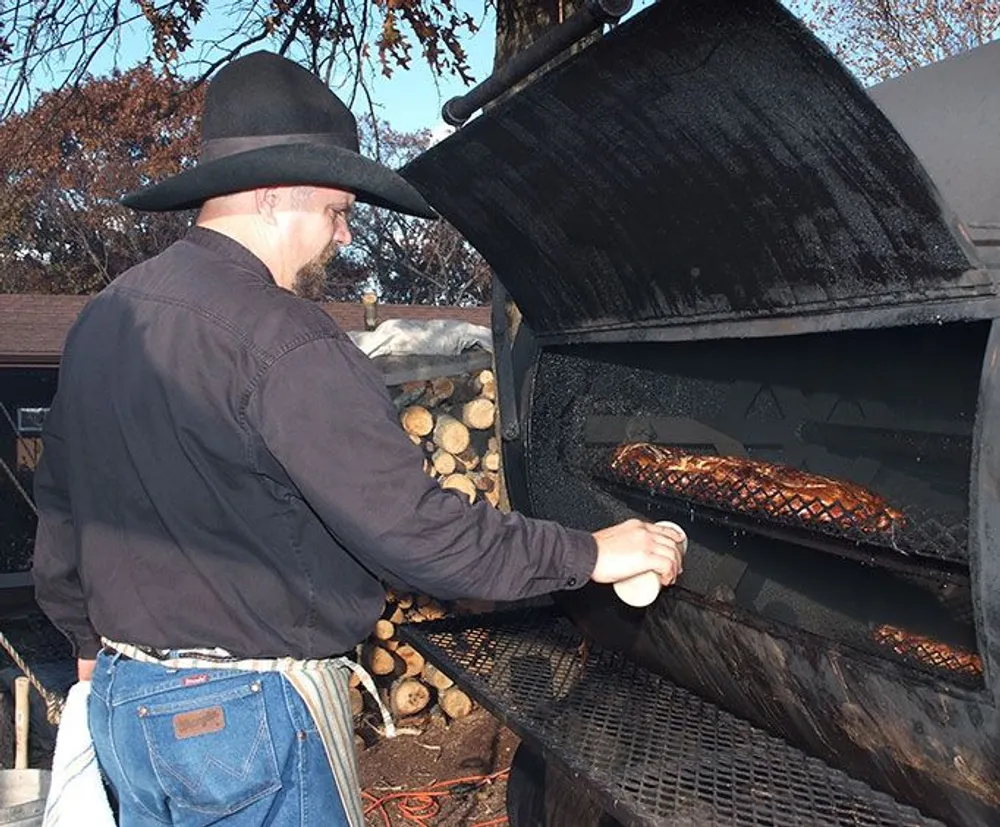 A man wearing a cowboy hat is tending to meat on a large outdoor barbecue smoker