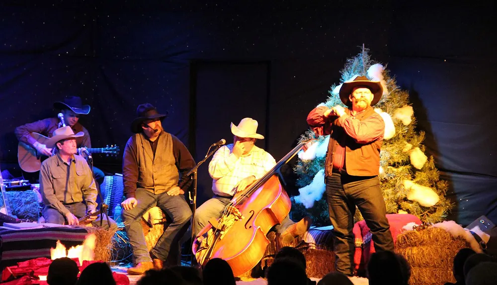 A group of men in cowboy hats perform music and storytelling on a festive stage with a Christmas tree