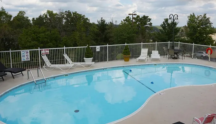 An outdoor swimming pool is surrounded by lounge chairs and a safety fence with a backdrop of trees and a cloudy sky