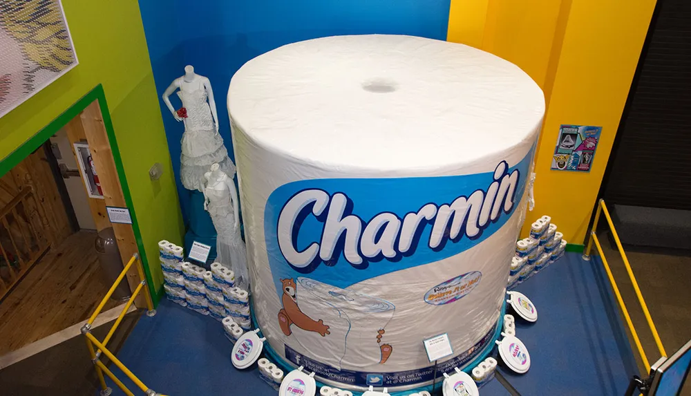 The image shows an oversized replica of a Charmin toilet paper roll in a colorful display room with a mannequin dressed in a wedding gown made of toilet paper to the left and several standard-sized products around