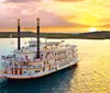 A paddle steamboat adorned with flags and bunting sails on a calm body of water during a vibrant sunset