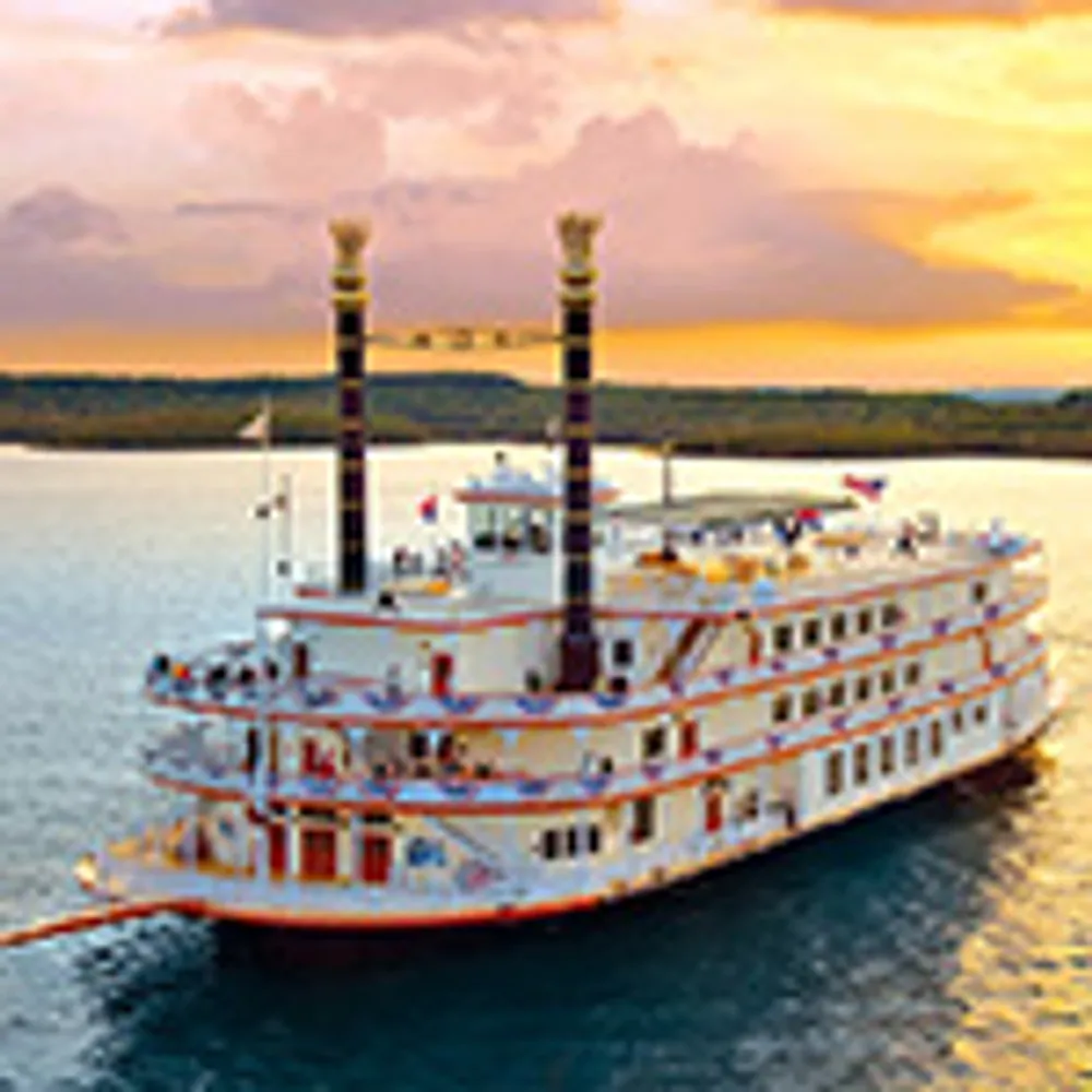 A white paddle steamer with two tall smokestacks is sailing on a calm body of water under a vibrant sunset sky