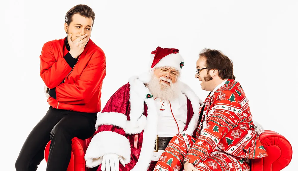 Three people are humorously posed in festive attire against a white background with an individual dressed as Santa Claus sitting between a person in a red tracksuit looking mockingly surprised and another in a vibrant Christmas suit whispering into Santas ear