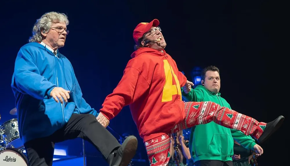Three men in colorful hoodies and Christmas-themed pants are performing energetically on stage