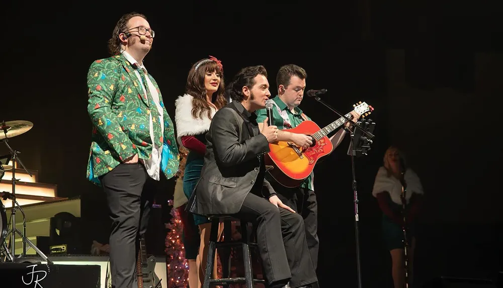 A group of performers dressed in festive attire are on stage with one person playing an acoustic guitar while another sings into a microphone