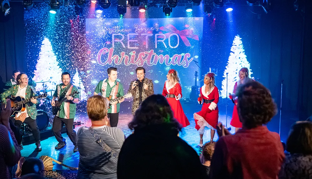 A retro Christmas-themed concert with performers in festive attire entertains a lively audience set against a backdrop of white Christmas trees and colorful lights