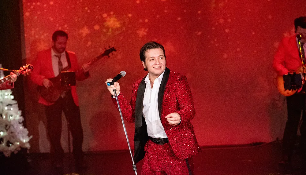 A performer in a red sequined suit sings into a microphone on stage accompanied by musicians in red jackets