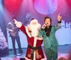 A performer in a sparkly suit is singing on stage alongside someone dressed as Santa Claus with a festive backdrop and musicians playing in the background