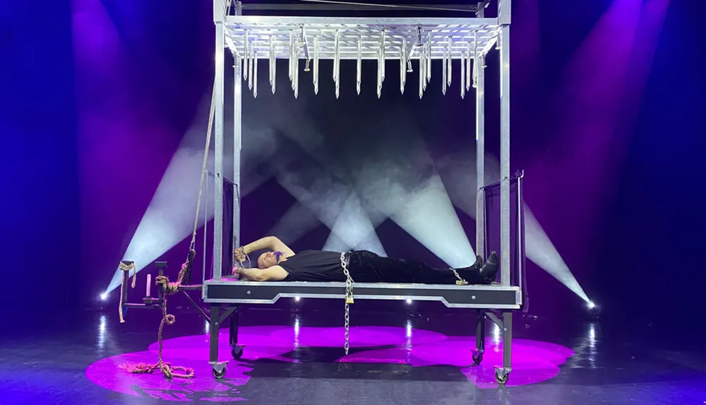 A person is lying on a table with chains on their wrists and ankles under a bed of suspended spikes illuminated by stage lights and enveloped in fog creating a dramatic escape artist scene
