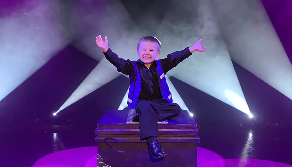 A child dressed in a dark outfit sits triumphantly atop a box on a stage with dramatic lighting radiating from behind