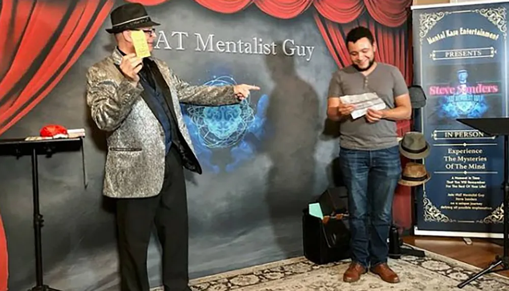 A person in a shiny silver jacket and hat is performing on stage pointing at a smiling audience member who is reading something both surrounded by mentalism-themed decor